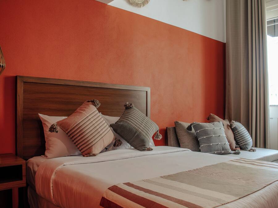 A hotel room with a wooden headboard, a bed with multiple pillows, a bedside table, and a window with curtains.