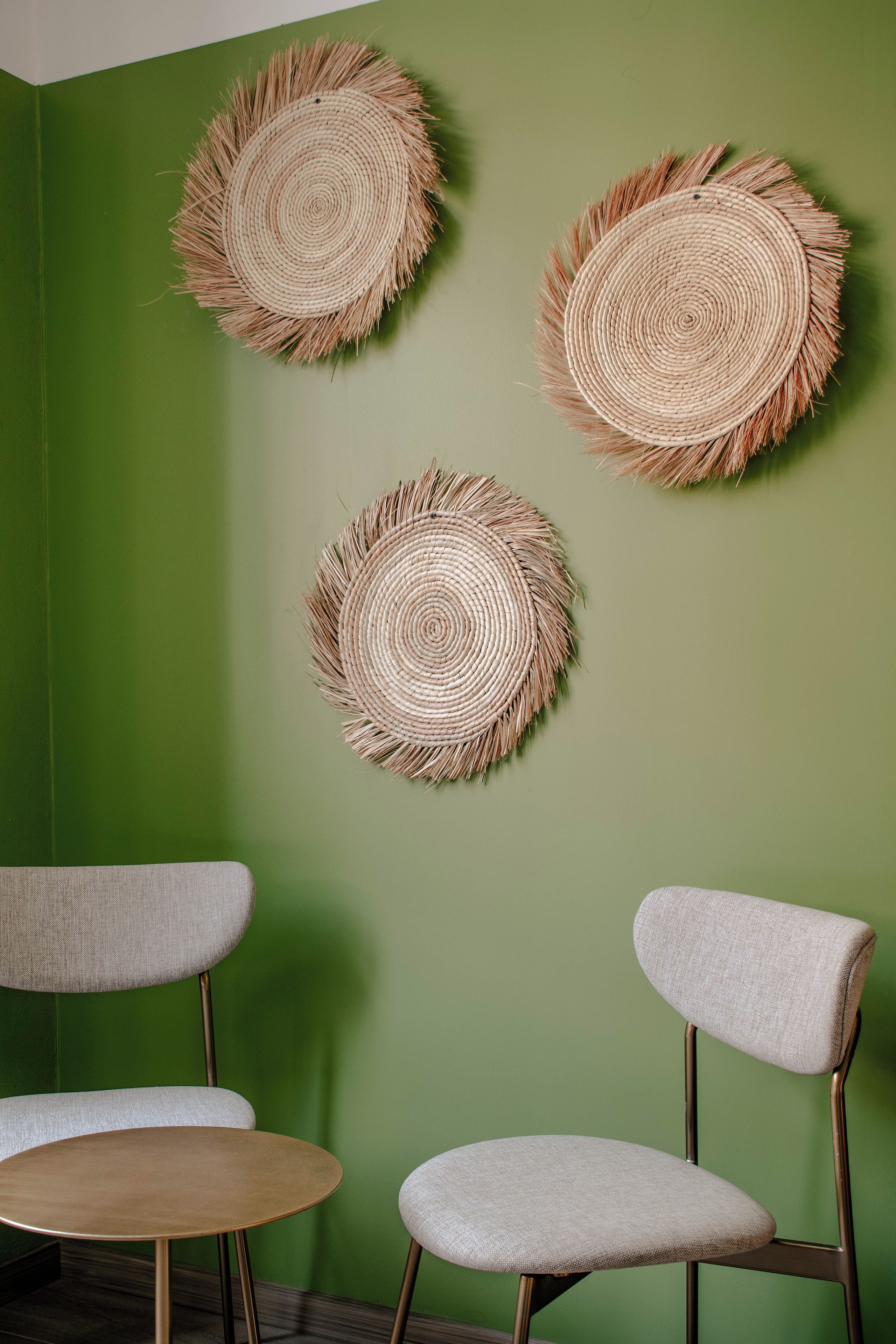 Una habitación con una pared verde y tres tapices de pared tejidos. Hay dos sillas con patas de metal y una mesa redonda.