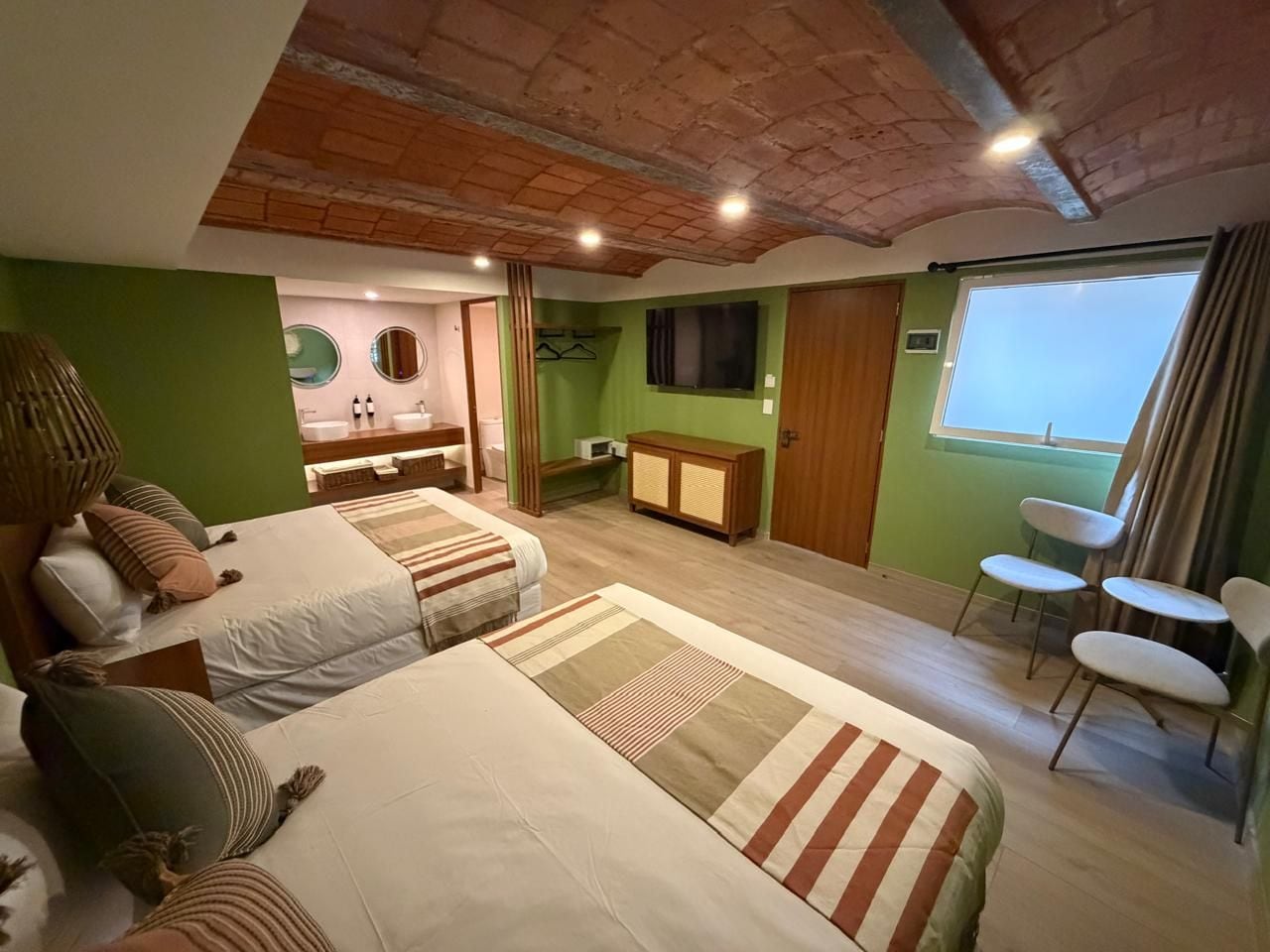 Una habitación con dos camas, una kitchenette pequeña, un ventanal con cortinas y una zona de estar con sillas.