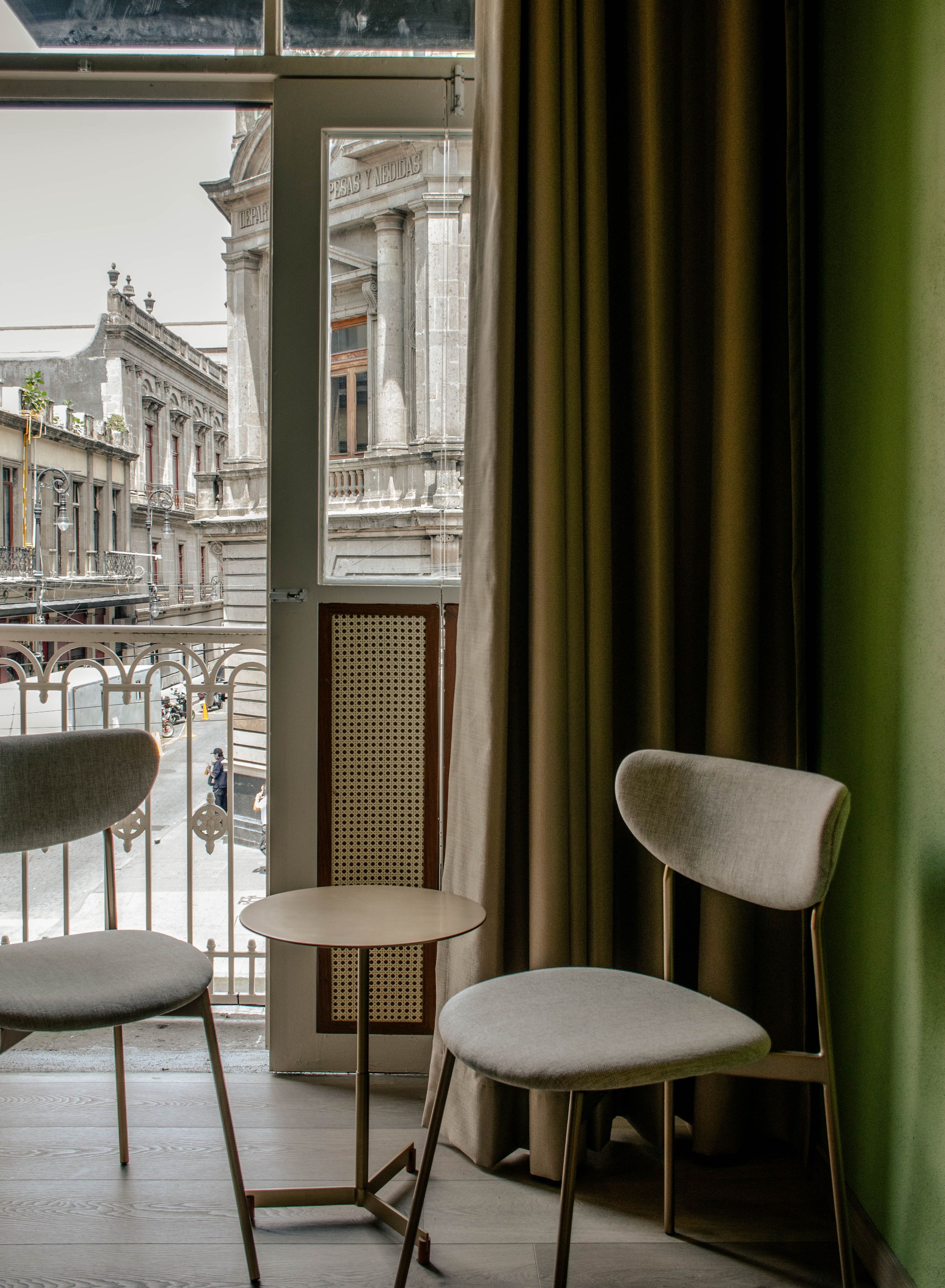 A room with two chairs and a small table, a window with a view of a building with columns, and a green curtain.