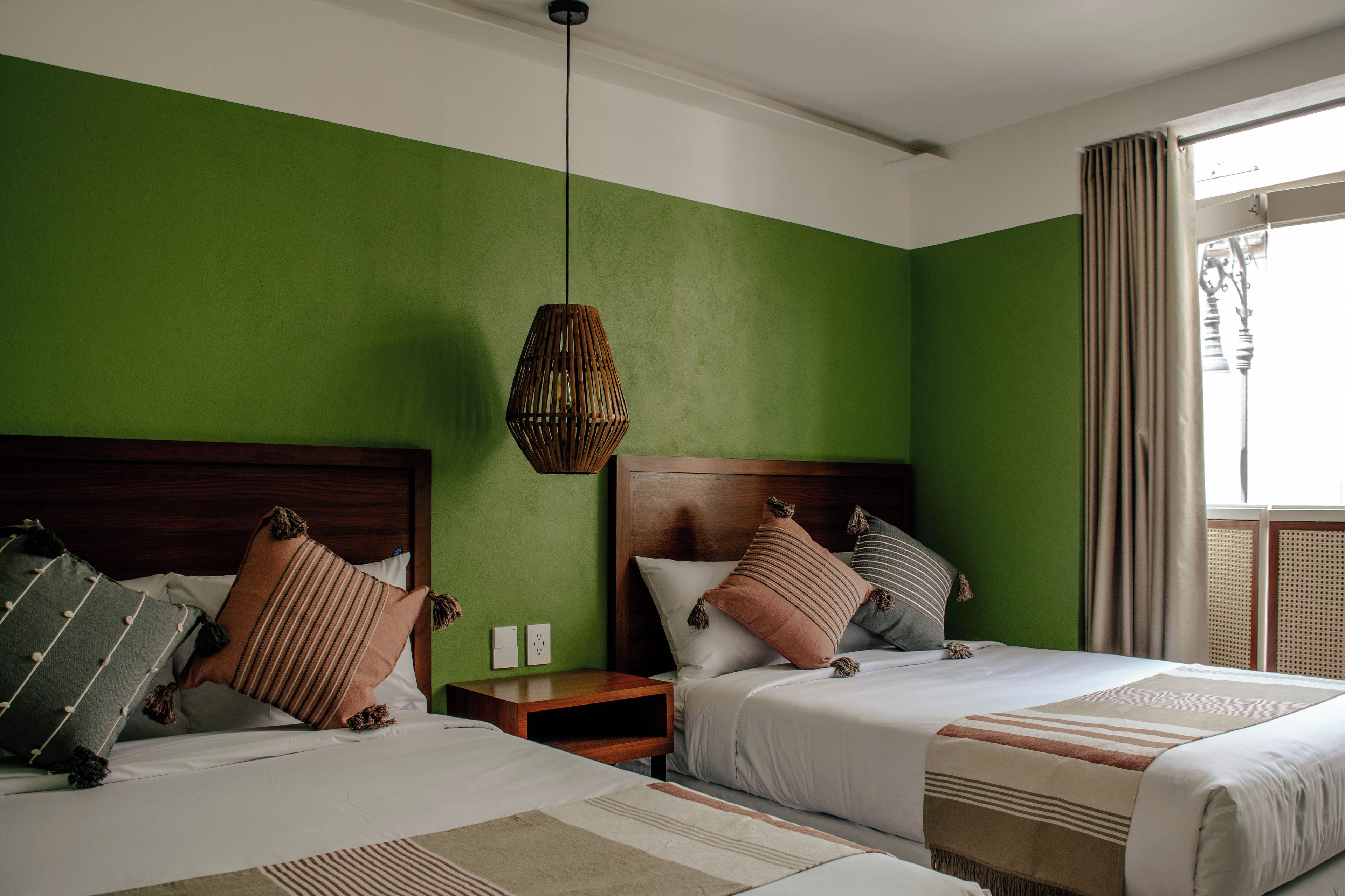 A hotel room with two beds, a green wall, a hanging light fixture, and a window with curtains.