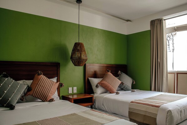 A hotel room with two beds, a green wall, a hanging light fixture, and a window with curtains.