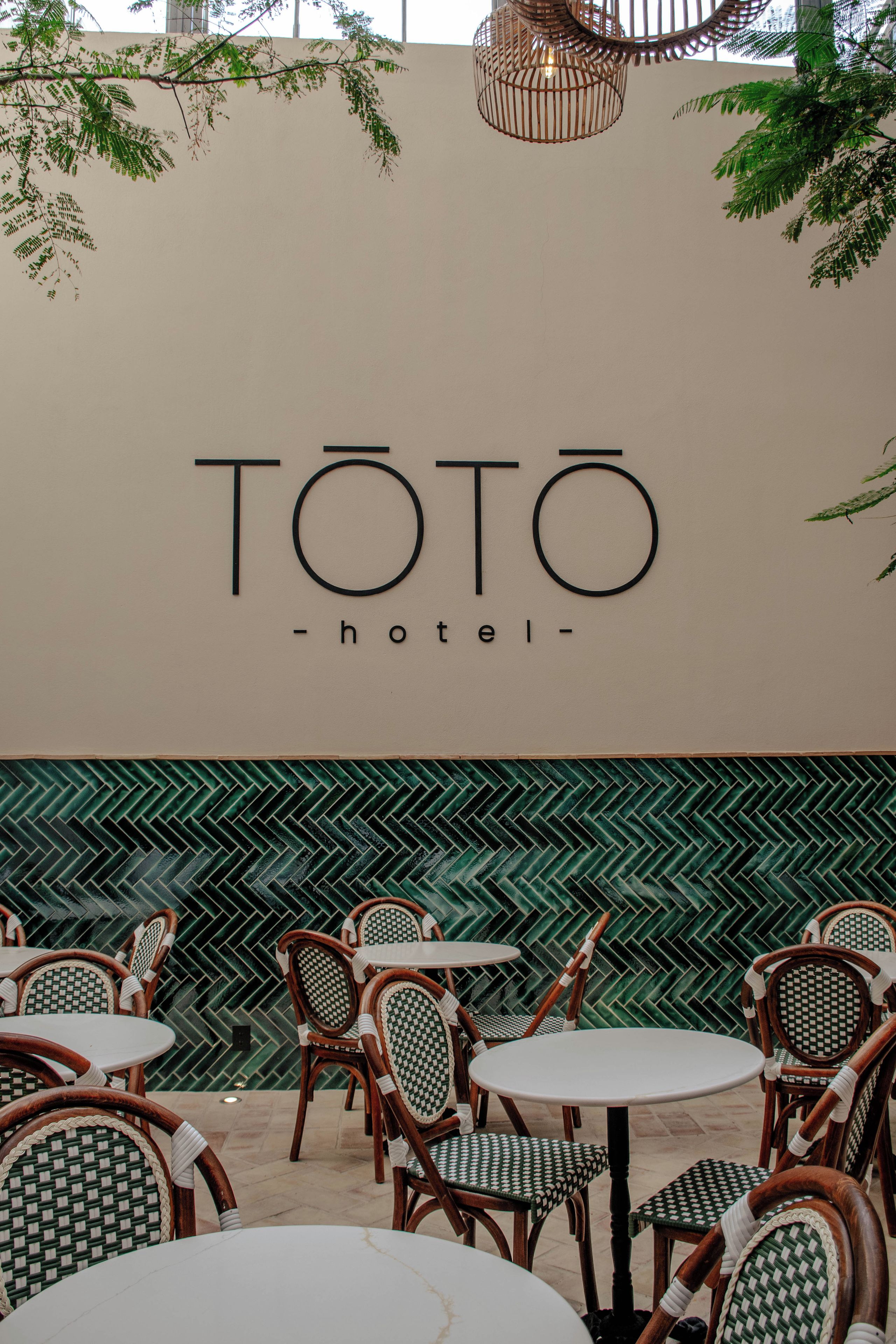 Un letrero del hotel TOTO con un diseño moderno, rodeado de asientos exteriores con sillas a cuadros y una pared con motivo de espiga.