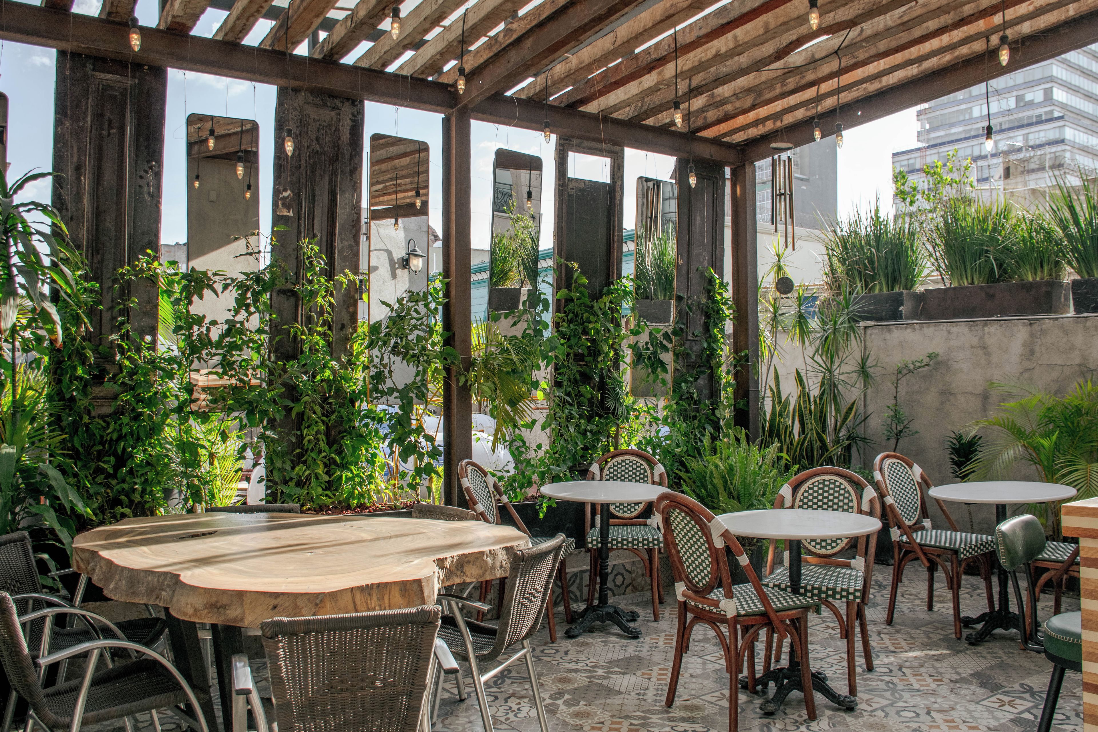 An outdoor patio with wooden tables and chairs, surrounded by lush greenery and a cityscape view.