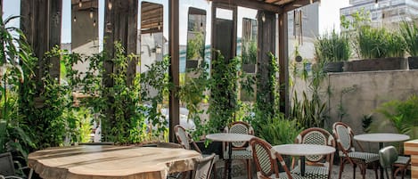 Un patio al aire libre con mesas y sillas de madera, rodeado de abundante vegetación y vistas a la ciudad.