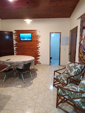 A room with a wooden ceiling, a round table with chairs, a television mounted on a wooden wall, and a colorful tapestry. - Beach house in Picinguaba, Beach house, House in Ubatuba, House in Picinguaba. (Ubatuba)