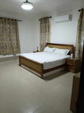 A bedroom with a wooden bed, white bedding, a nightstand, a wall-mounted air conditioner, and patterned curtains. - Luxurious Colonial Mansion in Puerto Plata With Private Pool (Puerto Plata)