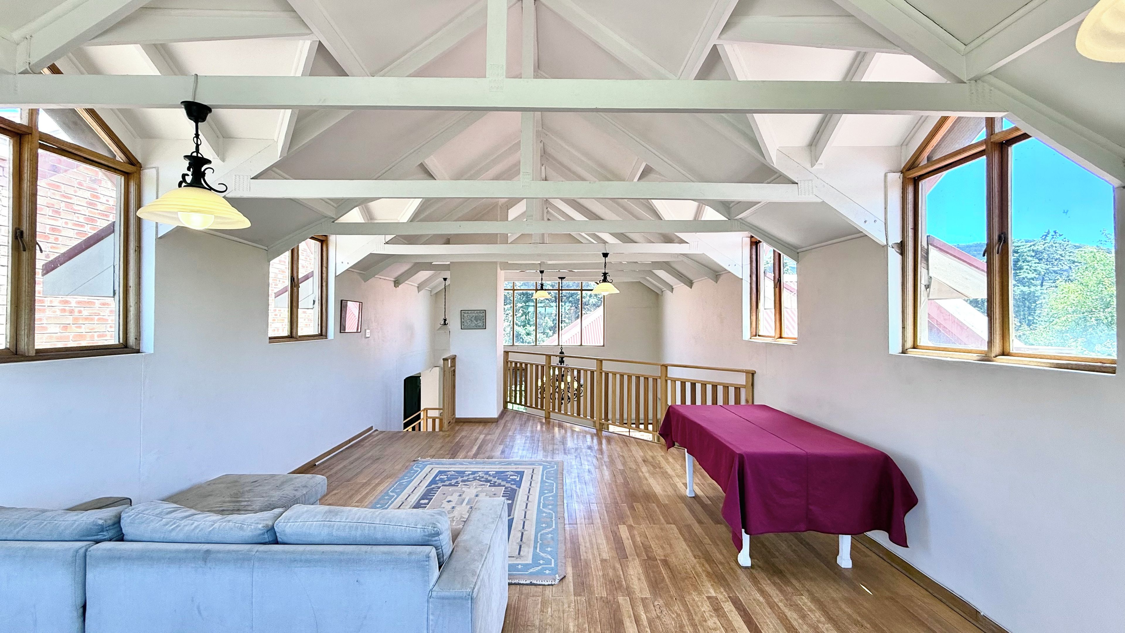 A spacious room with a high ceiling, wooden beams, a long table with a tablecloth, a sofa, and large windows offering a view of the outdoors.
