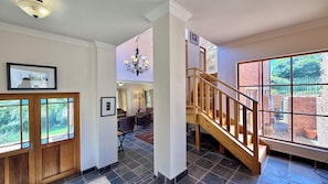 A spacious interior with a wooden staircase, a chandelier, and a view through large windows. - The Eyrie Guesthouse (Clarens)