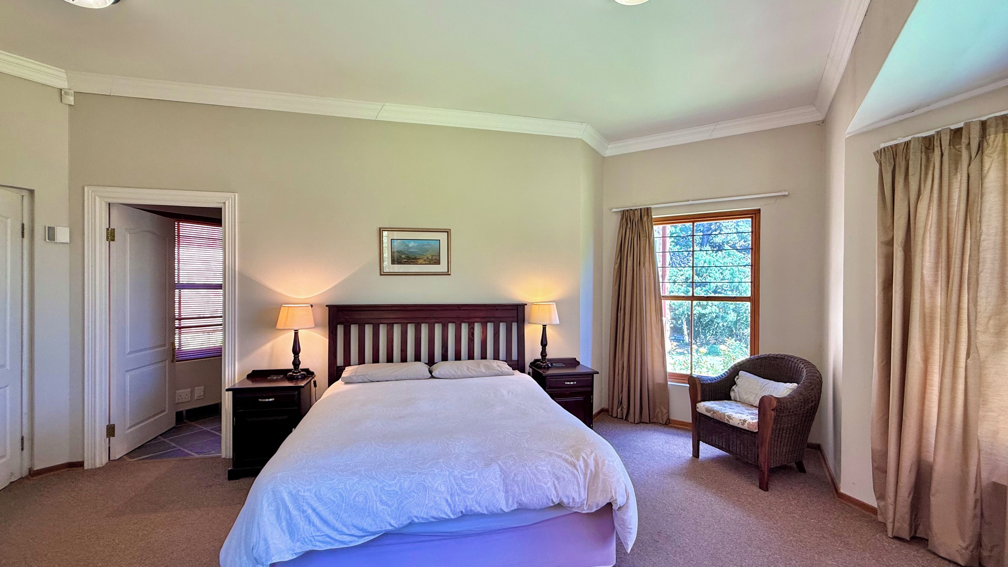 Una habitación de hotel con una cama grande, dos mesitas de noche con lámparas, una silla, una ventana con cortinas y un cuadro en la pared.