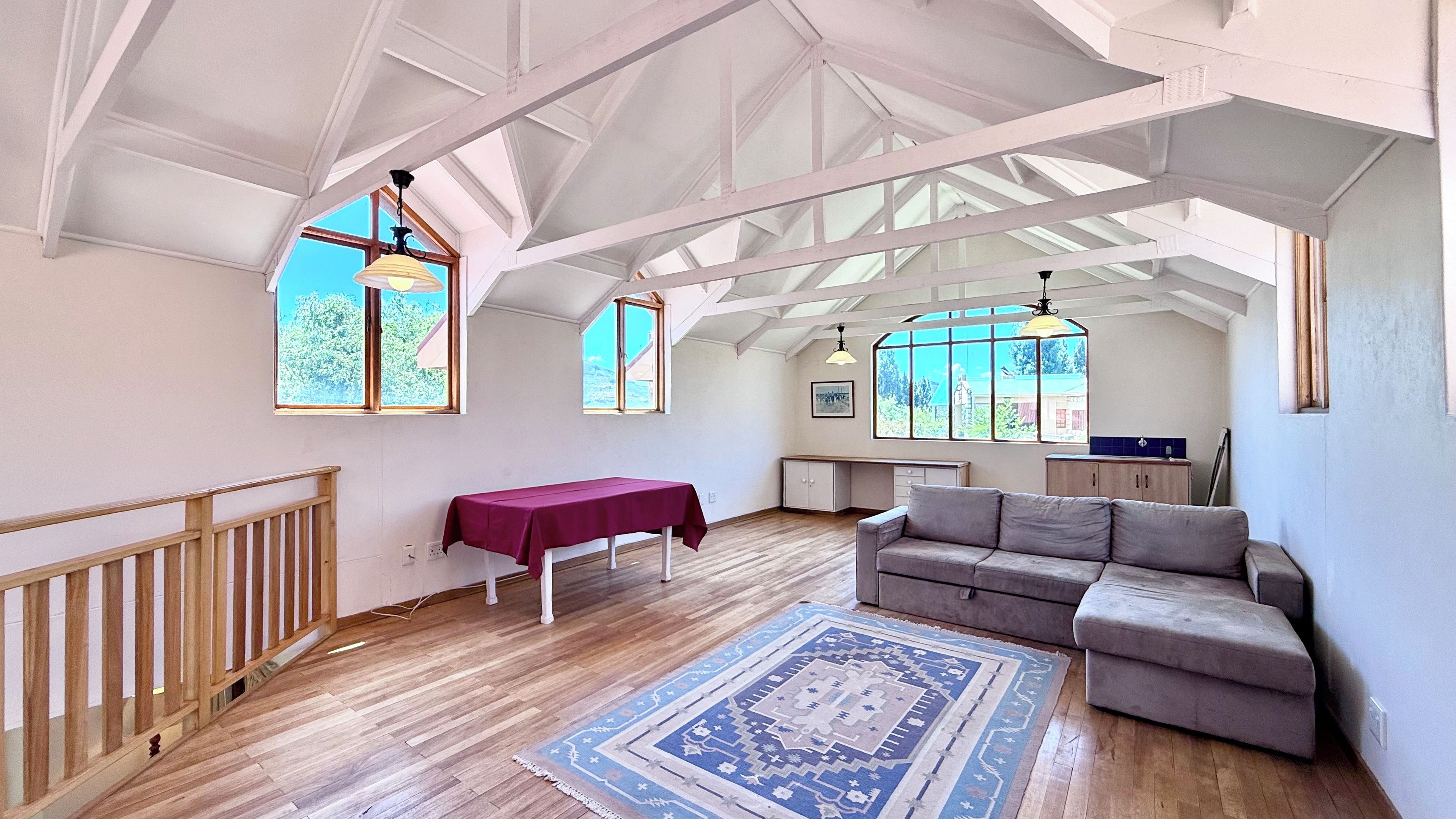 En rummelig stue med høje lofter, træbjælker, et spisebord dækket af en rød dug, en grå sofa og en tæppe med et mønster.