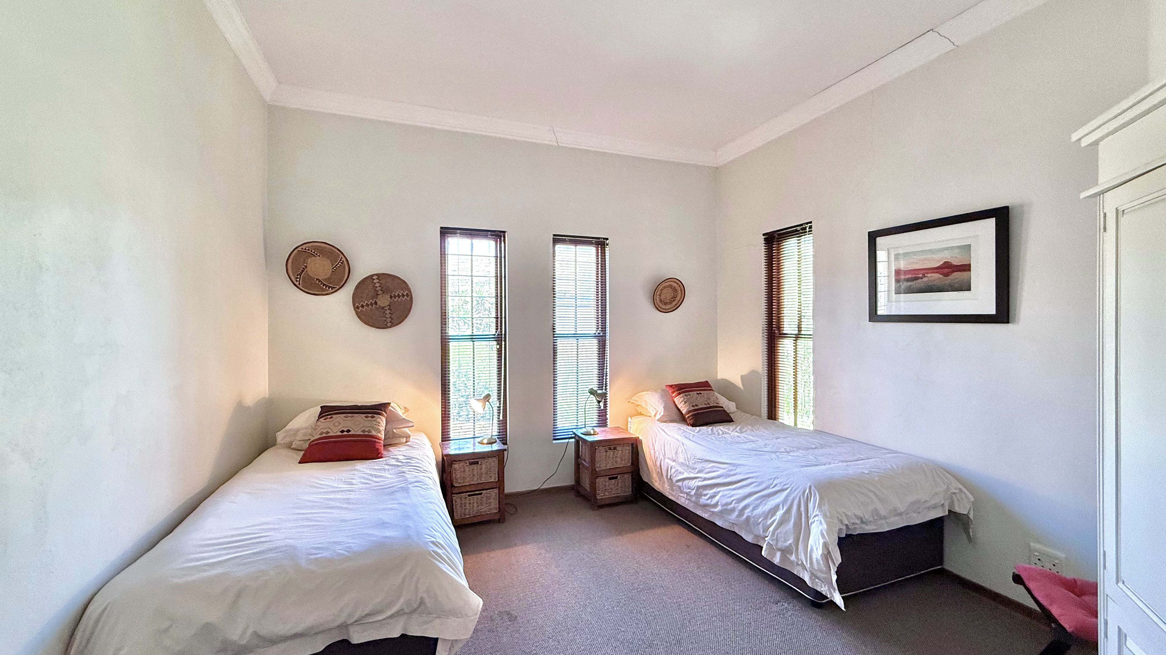 A room with two single beds, a framed picture on the wall, and two windows with blinds.