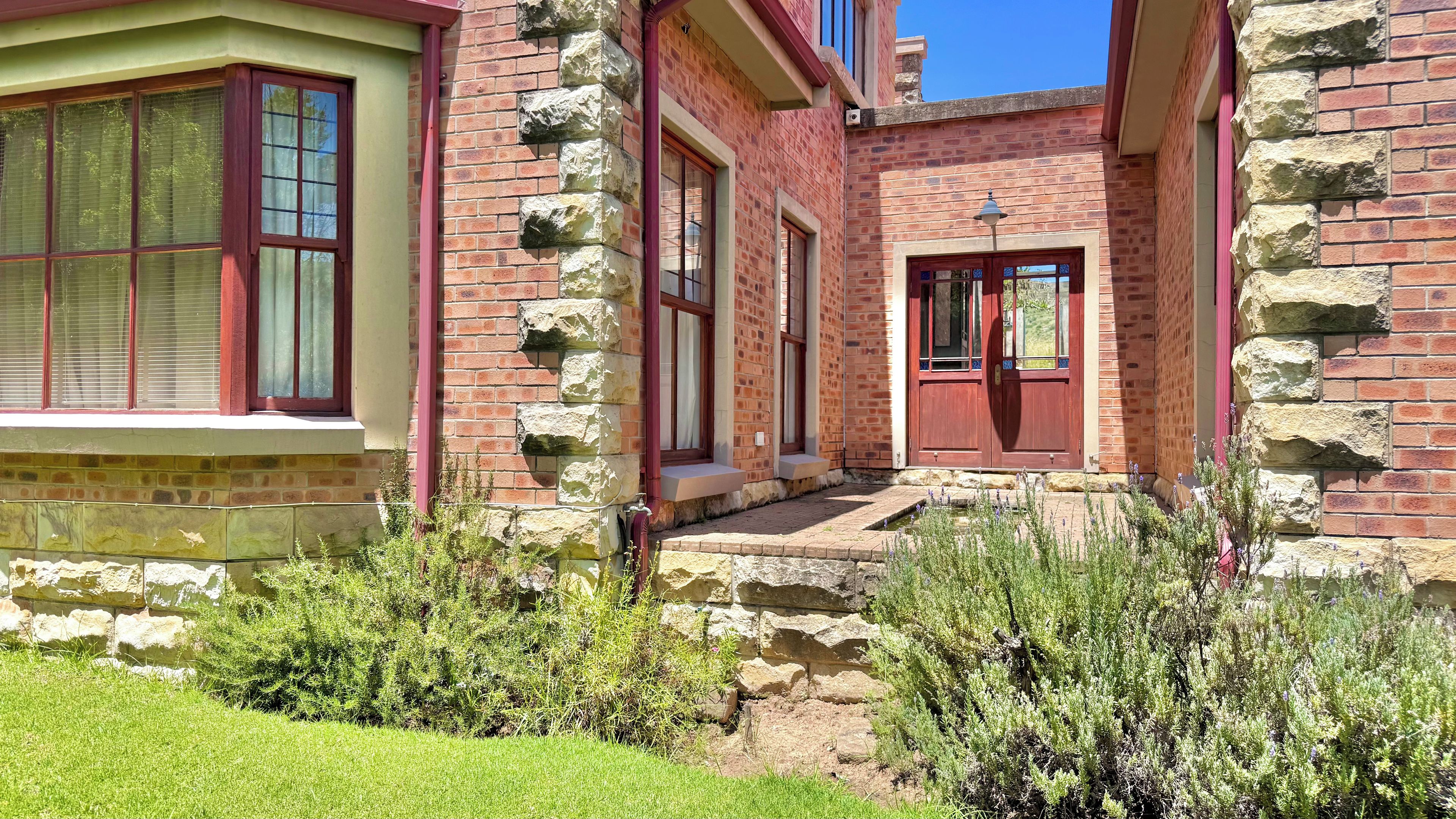 A stone building with a red door, windows with shutters, and a small garden area with plants.