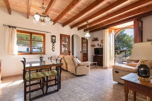A spacious living room with a dining area, wooden ceiling beams, a large window, and a view of the outdoors. - Es Turo Des Bessons by Rentallorca (Petra)