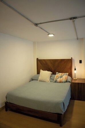 Une chambre à coucher avec une tête de lit en bois, un lit recouvert d’une courtepointe, deux oreillers, une table de chevet avec une lampe et un luminaire au plafond.