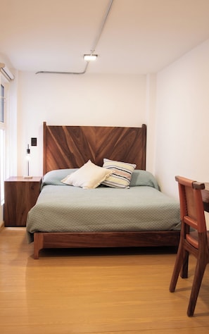 Een net opgemaakt bed met een houten hoofdbord, een nachtkastje met een lamp en een houten stoel.