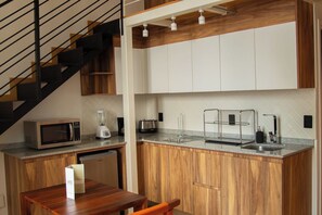 A compact kitchen with a microwave, toaster, and blender on the counter, wooden cabinets, and a staircase with black railings. - Loft histórico en el corazón de GDL LC (Guadalajara)