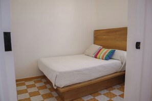 Een enkel bed met een houten hoofdbord en een kleurrijk gestreept kussen in een kamer met een geruit vloertje.