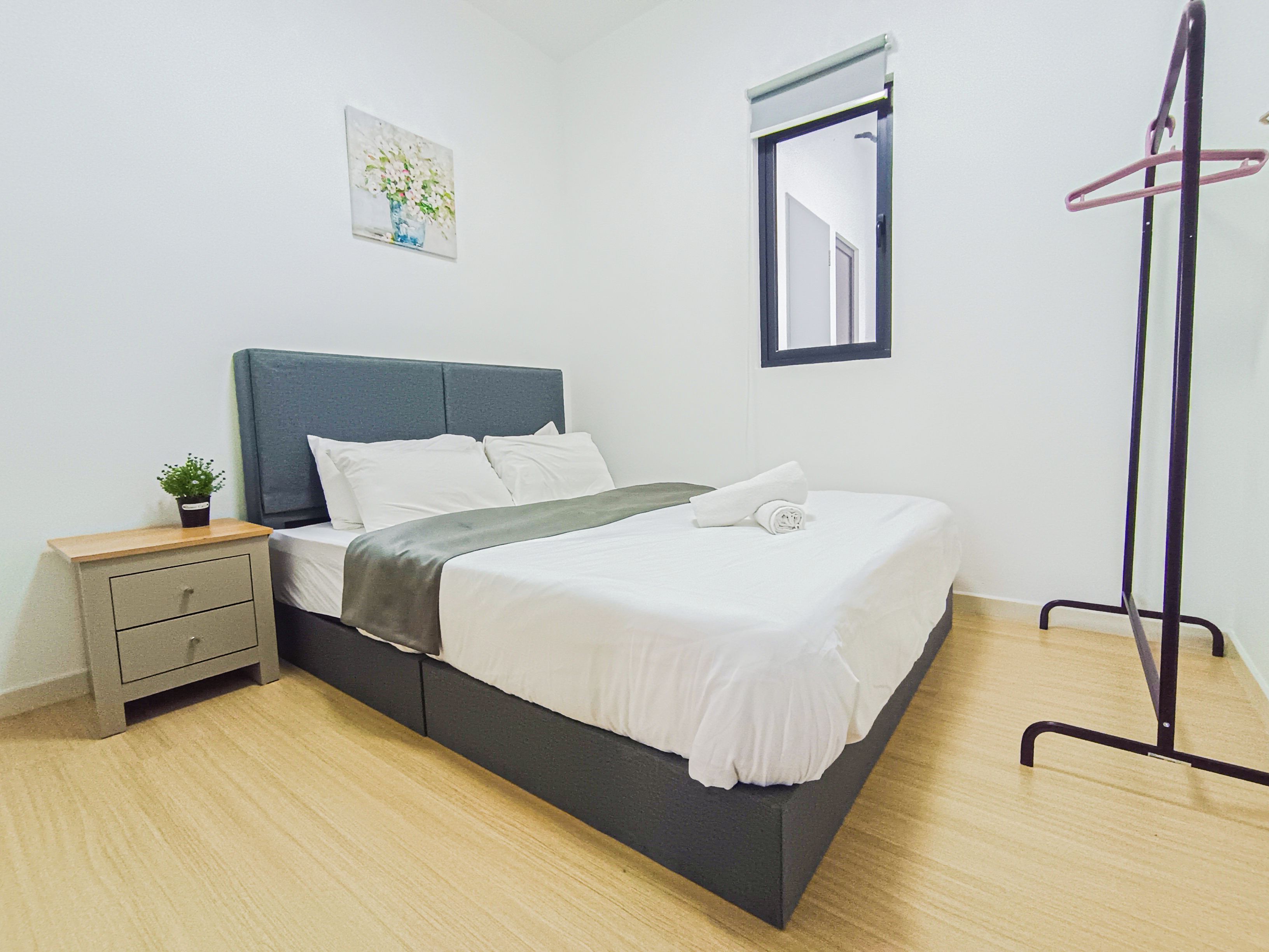 Een netjes opgemaakt bed met witte lakens en een grijs hoofdeinde, een nachtkastje met een plant, een klein schilderij aan de muur en een spiegel boven het bed.