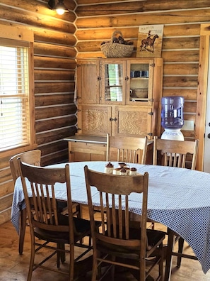 Interior de una cabaña de madera con una mesa de comedor, sillas, un aparador con puertas de cristal y un dispensador de agua.