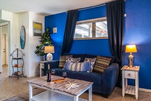 Ein Wohnzimmer mit blauer Wand, einem Sofa mit gemusterten Kissen, einem Couchtisch mit Snacks und einer Lampe auf einem Beistelltisch.