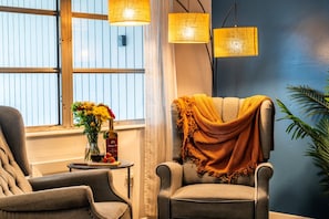 Ein Zimmer mit zwei Sesseln, einem kleinen Tisch mit Flasche und Blumen sowie einem Fenster mit Vorhängen.