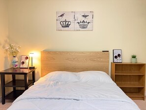 A bedroom with a wooden headboard, a bed with white bedding, a nightstand with a lamp, a framed picture, a vase with flowers, and a shelf with a plant. - Comfortable Basement Unit (Pickering)