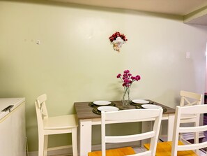 A dining area with a table set for four, white chairs, a vase with flowers, and a wall-mounted decoration. - Comfortable Basement Unit (Pickering)