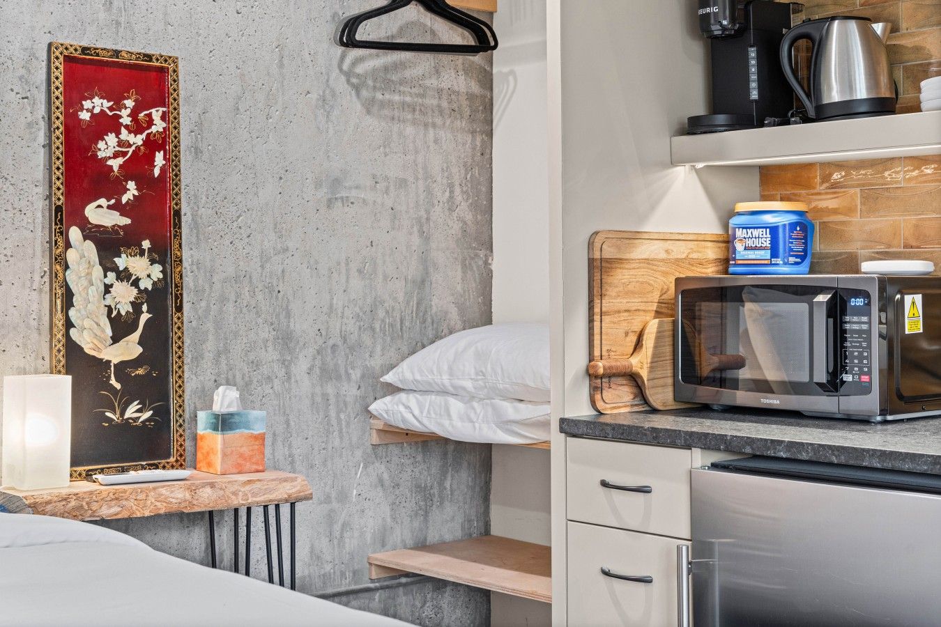 A hotel room with a bed, a bedside table with a lamp, a wall-mounted shelf with a kettle, a microwave, and a blue container.