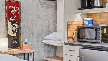 A hotel room with a bed, a bedside table with a lamp, a wall-mounted shelf with a kettle, a microwave, and a blue container.
