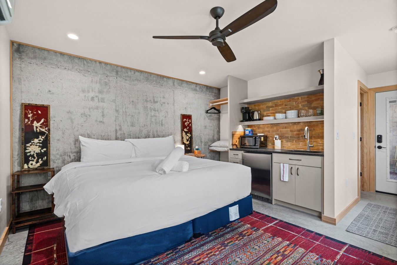 A hotel room with a bed, a ceiling fan, a small kitchenette, and a red patterned rug.
