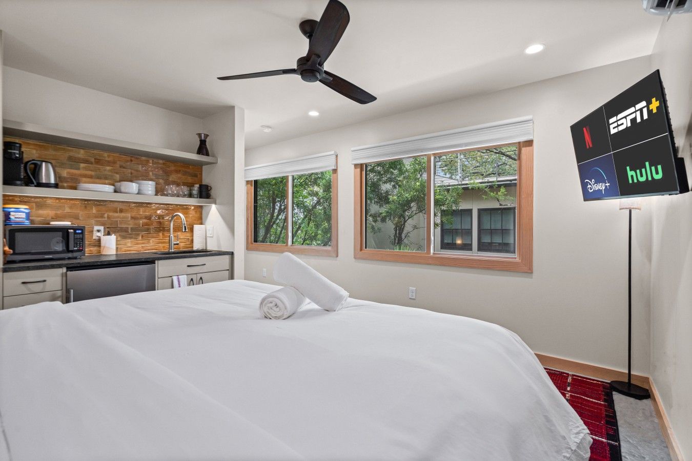 A hotel room with a bed, a ceiling fan, a kitchenette, and a TV displaying ESPN+ and Hulu logos.
