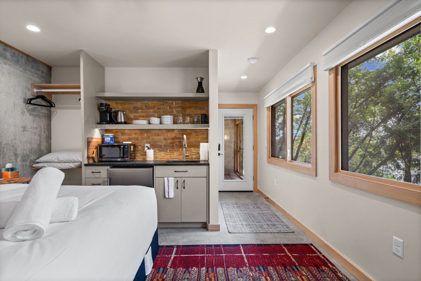 A modern bedroom with a bed, a small kitchenette, and a large window.