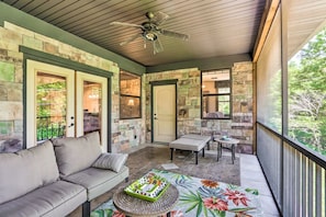 A covered patio area with a stone wall, a ceiling fan, a sofa, a table, and a view of greenery outside. - Dragon Castle: Secret Arcade, Hot Tub, Sleeps 22 (Camdenton)