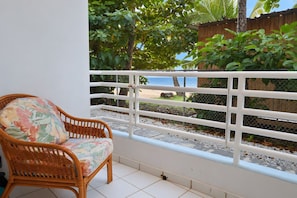 Una sedia in vimini con un cuscino a fiori, una ringhiera da balcone bianca e vista su una spiaggia e palme.