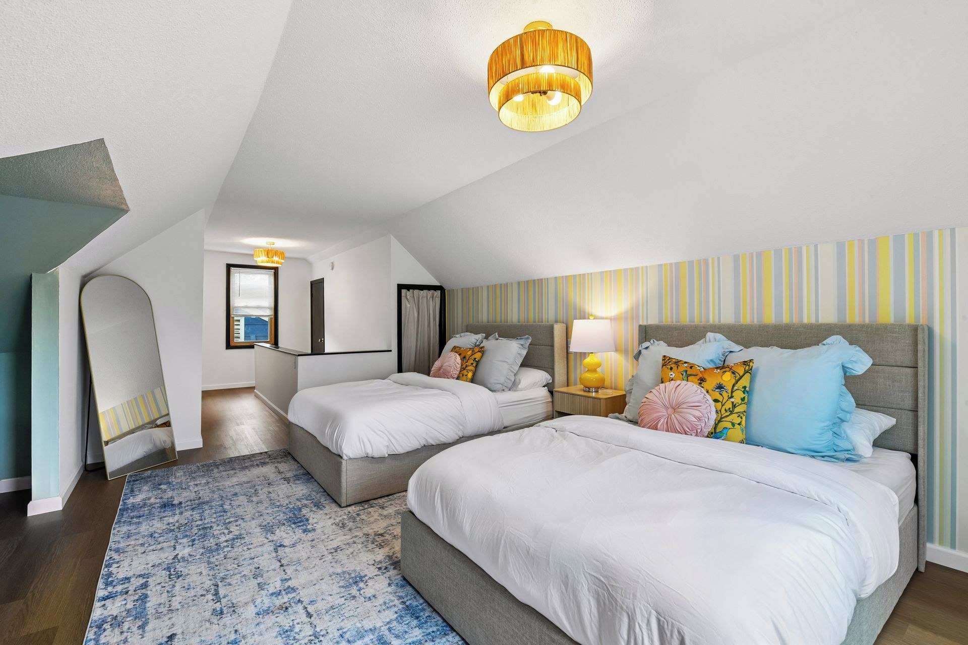 A modern hotel room with a sloped ceiling, two beds, a desk, a chair, a large window, and a striped wall.