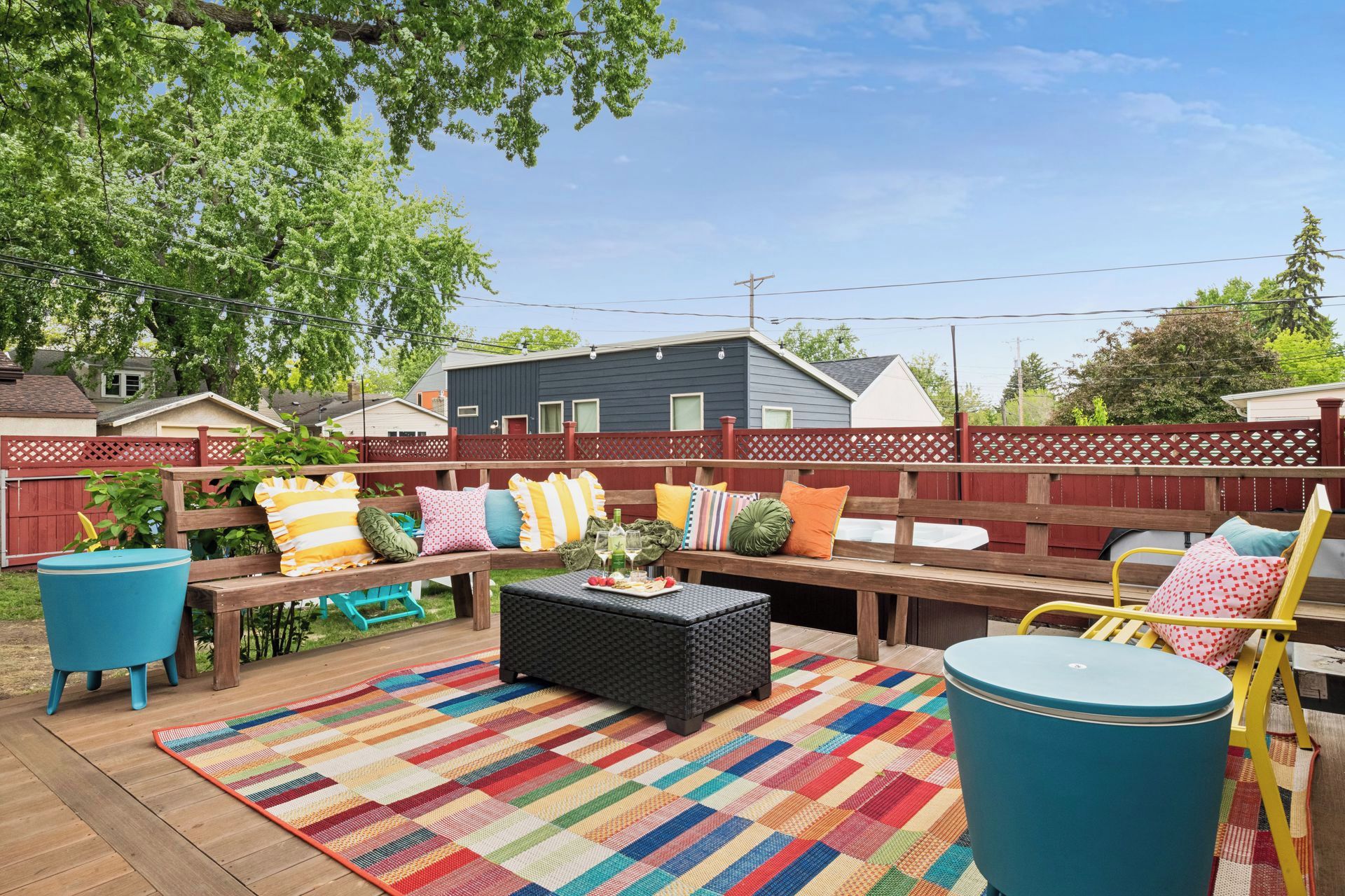 An outdoor patio with a wooden bench, colorful cushions, a woven coffee table, and a blue trash can.