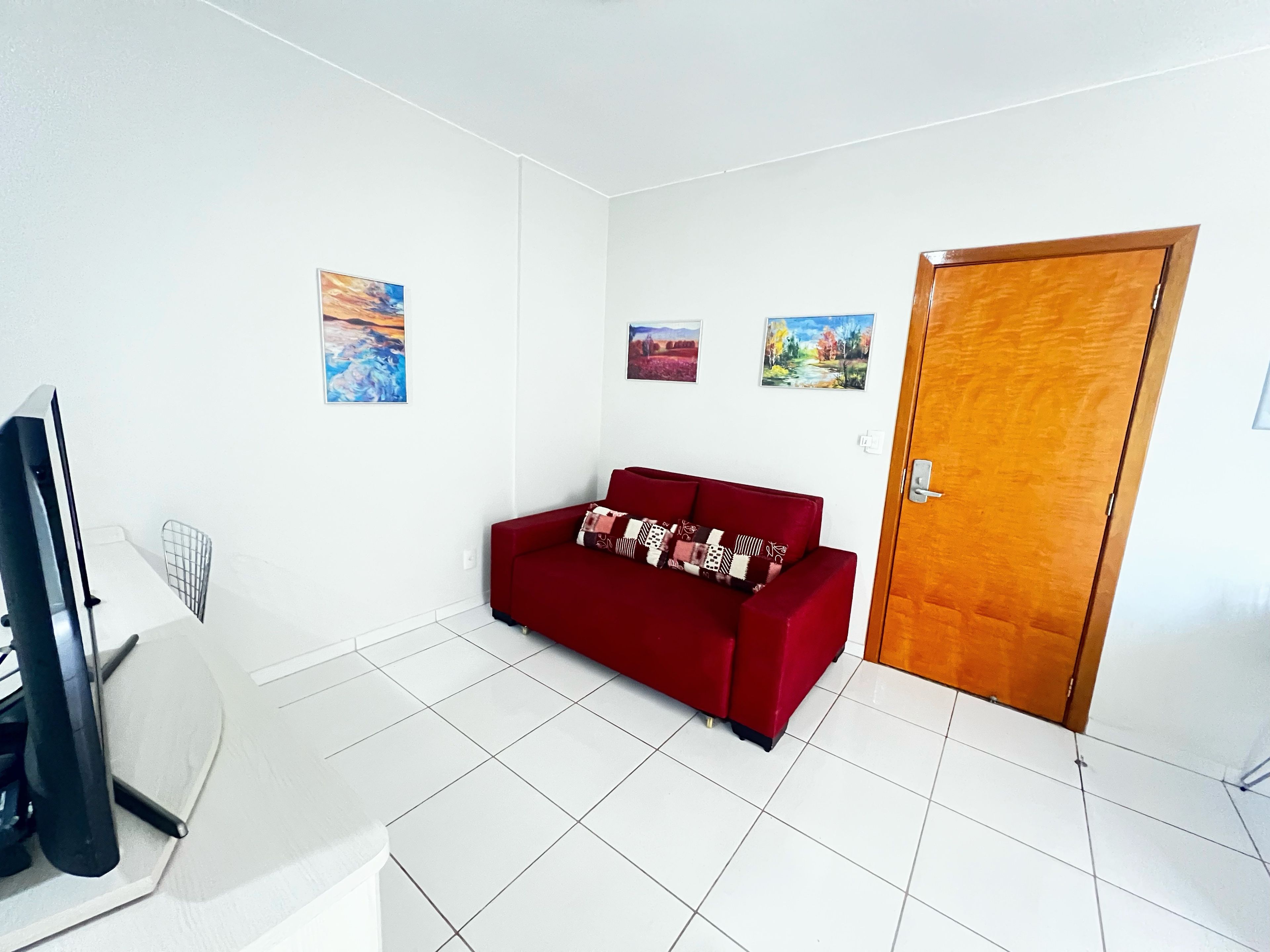 Una habitación con un sofá rojo, suelo de baldosas blancas, una puerta de madera y tres cuadros en la pared.