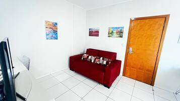 Una habitación con un sofá rojo, suelo de baldosas blancas, una puerta de madera y tres cuadros en la pared.