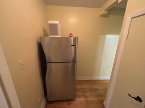 A stainless steel refrigerator in a narrow hallway with wooden flooring and a white door on the right.