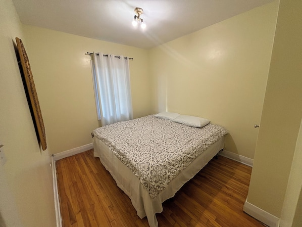 A bedroom with a single bed, a window with curtains, a wooden floor, and a ceiling light fixture.