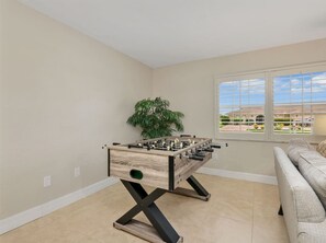 A room with a foosball table, a sofa, a potted plant, and a window with a view of a building. - Coral Cabana | Heated Pool | Fenced Yard | Bikes (Cape Coral)