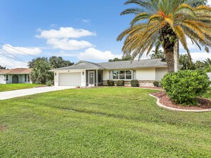 A single-story house with a well-maintained lawn, a palm tree, and a small bush. - Coral Cabana | Heated Pool | Fenced Yard | Bikes (Cape Coral)
