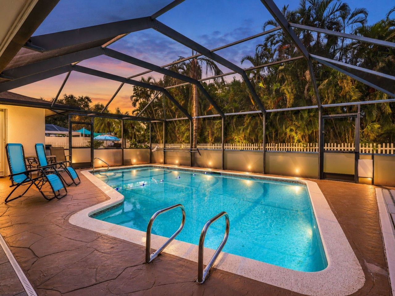Una piscina interior con techo de cristal, rodeada de palmeras y una valla de piquetes blancos.