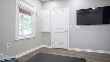 Ein Zimmer mit einem großen Flachbildfernseher an der Wand, einem Fenster mit Jalousien, einer Tür und einer dunkelgrauen Yogamatte auf dem Boden.