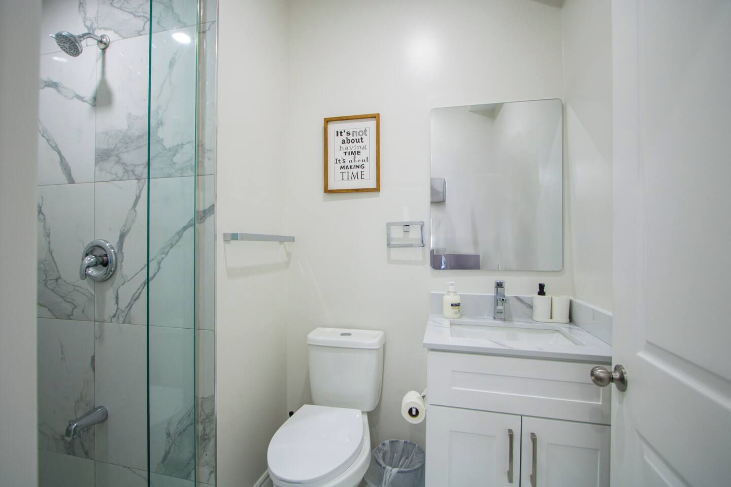 Un baño con cabina de ducha de vidrio, inodoro, lavamanos con espejo y una cita inspiradora enmarcada en la pared.
