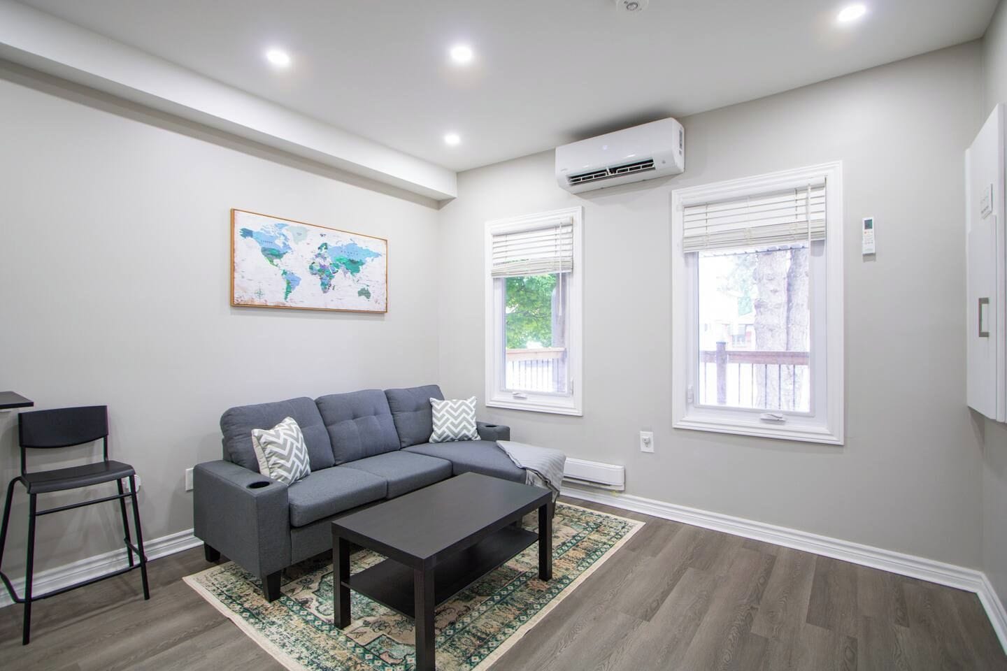 A modern living room with a grey sofa, a coffee table, a world map on the wall, and two windows with blinds.