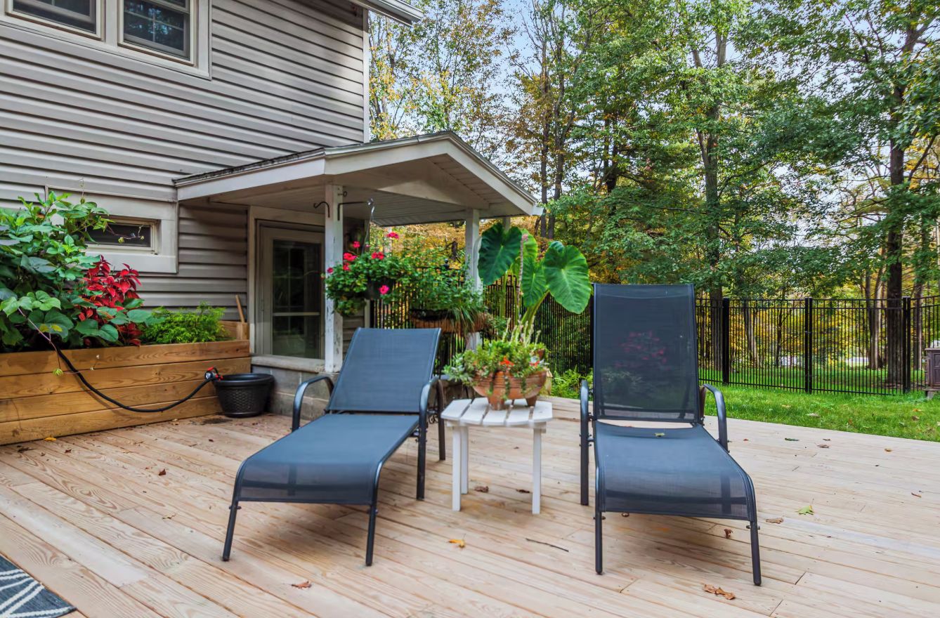 A covered outdoor deck with two lounge chairs and a small table, surrounded by plants and a wooden planter.