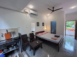 A compact hotel room with a bed, a desk, a chair, a small kitchenette, and a ceiling fan. - Studio Silent Grove R-6 Couple Choice (Arambol)