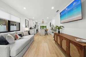 A modern living room with a grey sofa, a wooden console table, and a large TV screen displaying a beach scene. - The Cove in the Grove Bungalow in Historic Coconut (Miami)